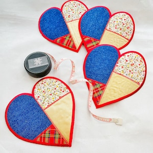 Op de afbeelding: Vier hartvormige stoffen onderzetters met een patchwork-ontwerp. Elke onderzetter heeft een blauwe denim stof, een crème stof met een bloemenprint, een crème stof met een rood-wit geruit patroon en een crème stof met een rood-witte bloemenprint.