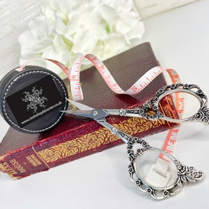 May include: A black leather tape measure with a white logo and a silver pair of decorative scissors with floral details. The tape measure is on top of a red book with gold lettering. The book is titled "Bleak House" by Charles Dickens.