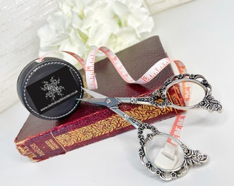 May include: A black leather tape measure with a white logo and a silver pair of decorative scissors with floral details. The tape measure is on top of a red book with gold lettering. The book is titled "Bleak House" by Charles Dickens.