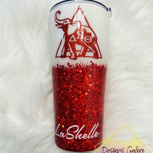 May include: A white and red glitter tumbler with a red elephant and triangle design. The text "Delta Sigma Theta" is inside the triangle. The text "LaShelle" is on the bottom of the tumbler.