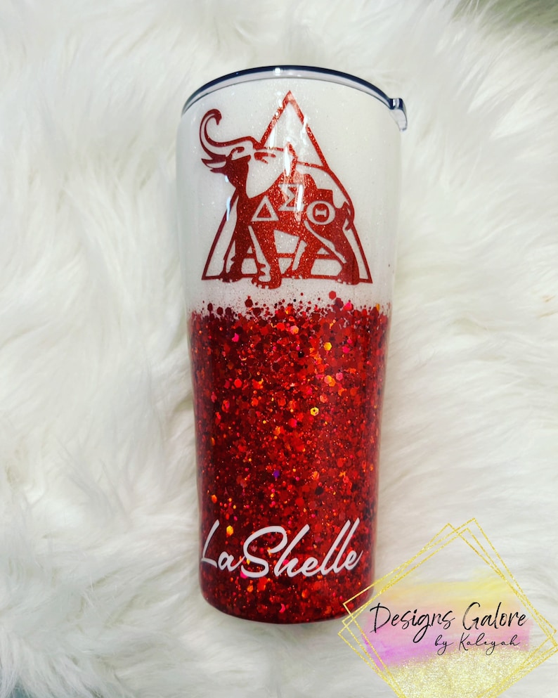May include: A white and red glitter tumbler with a red elephant and triangle design. The text "Delta Sigma Theta" is inside the triangle. The text "LaShelle" is on the bottom of the tumbler.