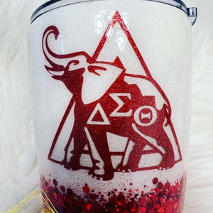 May include: A white and red glitter tumbler with a red elephant and the letters "&Delta;&Sigma;&Theta;" inside a triangle. The bottom of the tumbler is filled with red glitter.