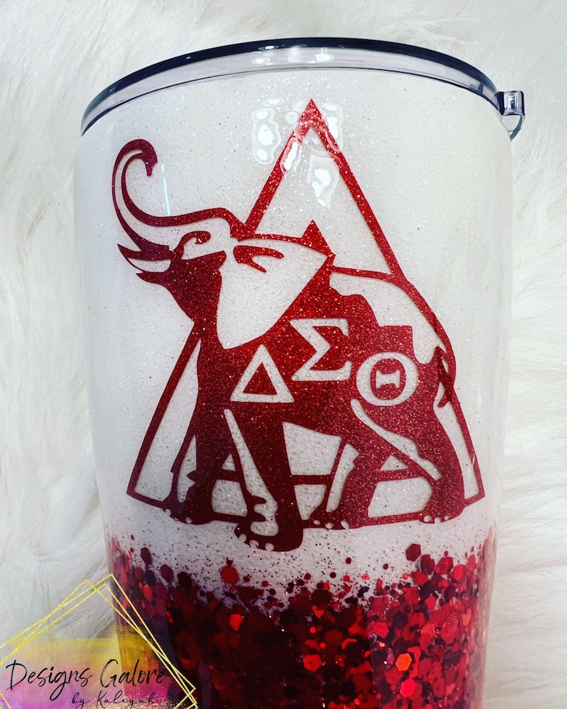 May include: A white and red glitter tumbler with a red elephant and the letters "&Delta;&Sigma;&Theta;" inside a triangle. The bottom of the tumbler is filled with red glitter.