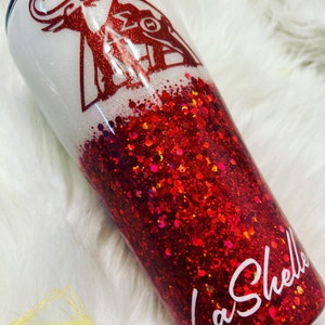 May include: A white and red glitter tumbler with a red elephant and Greek letters on the top half. The bottom half is red glitter. The tumbler has the name "La'Shelle" written in white on the bottom.
