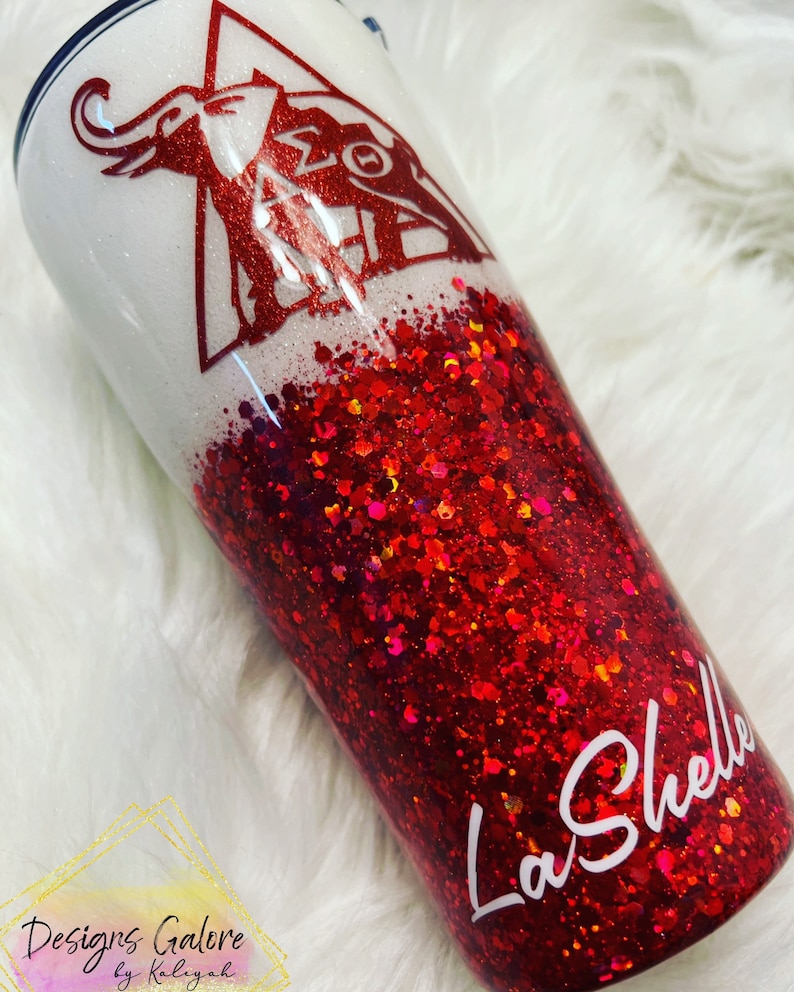 May include: A white and red glitter tumbler with a red elephant and Greek letters on the top half. The bottom half is red glitter. The tumbler has the name "La'Shelle" written in white on the bottom.
