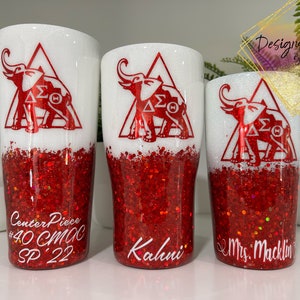 May include: Three white tumblers with red glitter bottoms. The tumblers have a red elephant design with the letters "&Delta;&Sigma;&Theta;" inside a triangle. The tumblers are labelled "Center Piece #40 CMC SP 22", "Kahui", and "Mrs. Macklin".