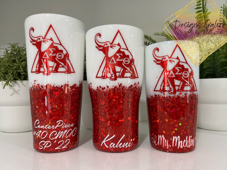 May include: Three white tumblers with red glitter bottoms. The tumblers have a red elephant design with the letters "&Delta;&Sigma;&Theta;" inside a triangle. The tumblers are labelled "Center Piece #40 CMC SP 22", "Kahui", and "Mrs. Macklin".