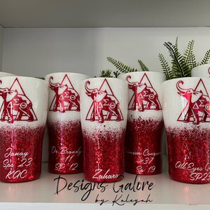 May include: Six red and white glitter tumblers with a white elephant and Greek letters in a triangle design. The tumblers have names and dates written on them.