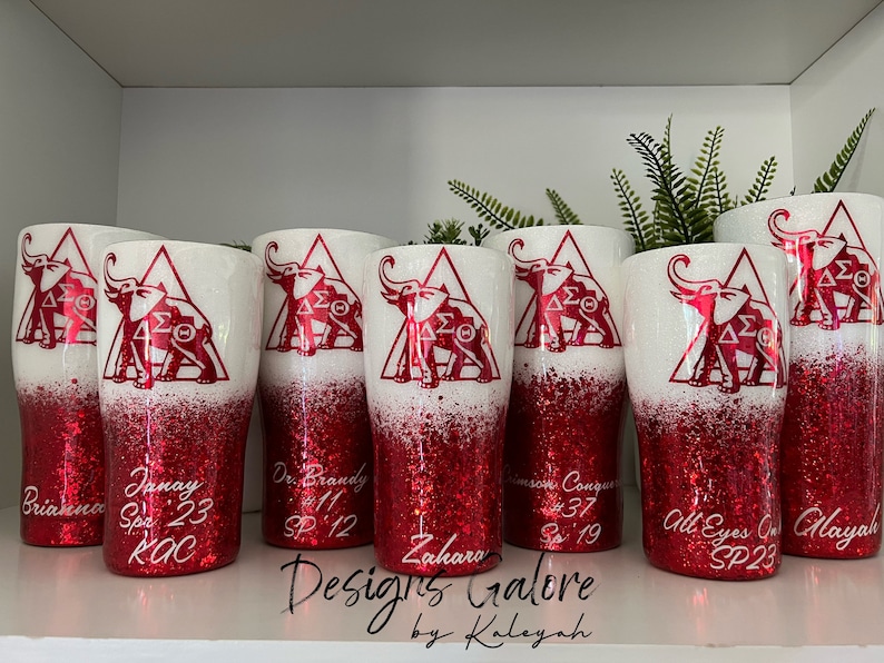 May include: Six red and white glitter tumblers with a white elephant and Greek letters in a triangle design. The tumblers have names and dates written on them.