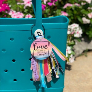 Pode incluir: Bolsa tote turquesa com um berloque decorativo. O berloque apresenta a palavra "love" e o versículo bíblico João 4:19, com fitas e borlas coloridas. A bolsa tem uma alça e um porta-berloque redondo.