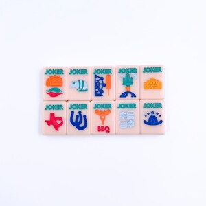 May include: A set of 12 pink dominoes with green, blue, orange, and red illustrations of Texas-themed icons. Each domino has the word "JOKER" printed on it.