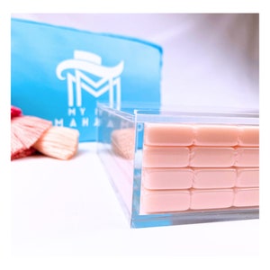 May include: A clear acrylic box filled with rows of light pink rectangular objects. A light blue bag with white text and a logo is in the background, along with a pink makeup brush.