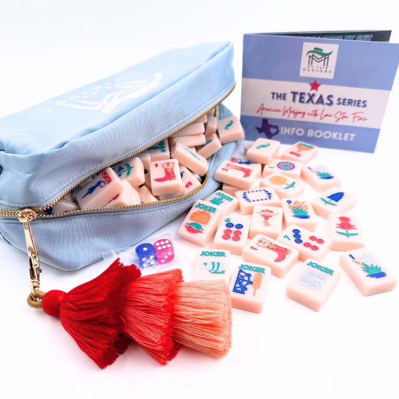 May include: A light blue zippered pouch with a white design and a tassel. The pouch is filled with a set of pink dominoes with colorful Texas-themed illustrations. The dominoes are scattered around the pouch, along with two dice. The text "THE TEXAS SERIES" and "INFO BOOKLET" is visible on a blue booklet.