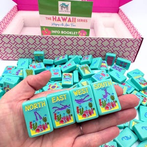 May include: A hand holds four turquoise mahjong tiles with colorful illustrations of underwater scenes. The tiles are labeled "North", "East", "West", and "South".