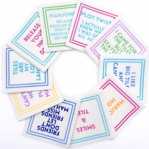 May include: A collection of white square tiles with colorful borders and text. The tiles feature phrases like "Plot Twist," "I Like Big Tiles," and "Friends Don't Let Friends Miss Mahjong." The tiles are arranged in a circular pattern.