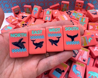 May include: A collection of coral-colored Mahjong tiles. The tiles feature illustrations of whales, palm trees, and other designs. The tiles are labeled with the cardinal directions: North, East, West, and South. The text and illustrations are in teal.