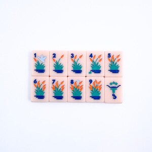May include: A set of nine pink mahjong tiles with blue and orange designs. The tiles are numbered 1 through 9 and feature a bird and a bull.