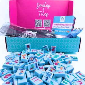 May include: A blue box with the words "Smiles & Tiles" on the lid. The box is open and contains a set of blue mahjong tiles with colorful designs. The tiles are scattered on a white surface. The box also contains a small booklet with the words "The Anywhere Series" and "Info Booklet".