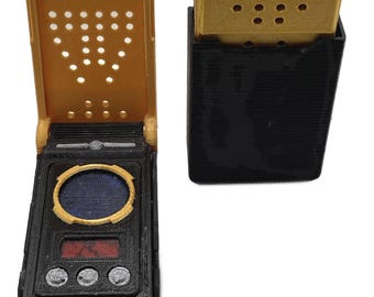 Könnte beinhalten: Zwei rechteckige Kommunikationsgeräte mit 3D-Druck-Struktur. Ein Gerät ist geöffnet und zeigt einen goldenen Deckel mit Perforationen, ein blaues kreisförmiges Display, einen roten rechteckigen Bildschirm und drei silberne runde Tasten auf schwarzer Basis.