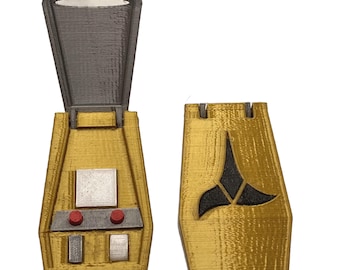 Könnte beinhalten: Ein 3D-gedrucktes goldenes Kommunikator-Requisit mit Klappdeckel. Die geöffnete Einheit zeigt ein graues Inneres mit einem silbernen Quadrat, zwei kleinen roten runden Knöpfen und zwei rechteckigen silbernen Knöpfen. Die geschlossene Einheit zeigt ein schwarzes deltaförmiges Emblem.