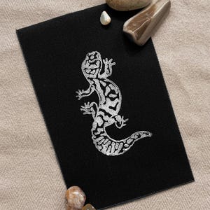 May include: A black fabric patch with a white embroidered image of a salamander. The salamander is facing right and has a long tail.