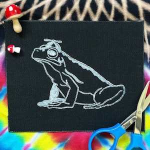 May include: A black square patch featuring a silver outline of a frog wearing a hat. Two small mushroom-shaped pins are placed near the patch. Blue-handled scissors are in the lower right corner. The background is a tie-dye pattern.