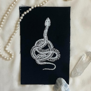 May include: A black fabric patch featuring a white, stylized snake skeleton design. A pearl necklace and two crystals are arranged near the patch on a white surface. The snake design is the focal point.