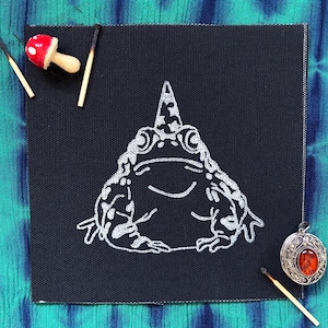 May include: A black square canvas with a white illustration of a smiling frog wearing a patterned hat. A red and white mushroom, a silver pendant with an orange stone, and burnt matches are arranged around the canvas on a tie-dye background.