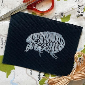 May include: A black fabric square with a white embroidered image of a flea. The fabric is on a white surface with a map and a pair of red scissors.