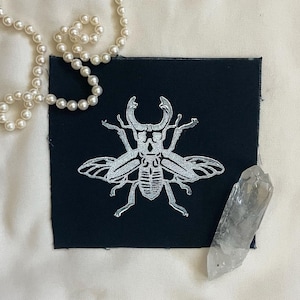 May include: A black fabric patch with a silver-colored stag beetle design. The beetle is detailed with intricate lines and has large, prominent antlers.