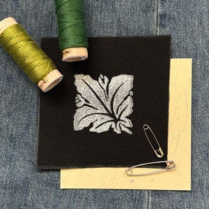 May include: A black square of fabric with a silver leaf design, two spools of green thread, and two silver safety pins. The fabric is set on a denim background. The thread spools are labeled "Italy".