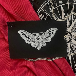 May include: A black patch with a white embroidered moth design. The moth has a skull and crossbones pattern on its body.