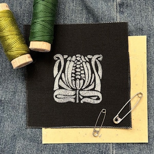 May include: A black fabric square with a white floral design, two spools of thread, two safety pins, and a piece of tan paper. The spools are green and yellow-green. The background is denim.