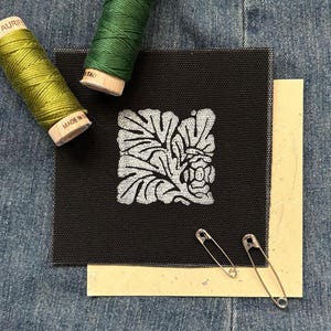 May include: A black square fabric with a white floral design, two safety pins, and two spools of thread in green and yellow-green. The fabric is layered over a piece of cream-colored paper, all on a denim background.