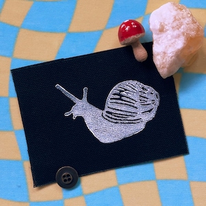 May include: A black fabric patch with a white embroidered snail design. The snail is facing left and has a detailed shell.