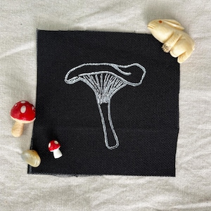 May include: A square black fabric patch featuring a white embroidered chanterelle mushroom. The patch is surrounded by two small red and white spotted mushroom figurines, a smooth tan stone, and a small carved cream-colored rabbit figurine.
