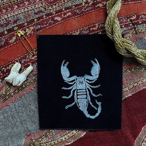 May include: A black fabric patch with a white embroidered scorpion design. The scorpion is facing right and has a detailed body and legs.