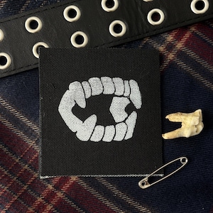 May include: A square black fabric patch featuring an embroidered white graphic of a set of vampire fangs. The patch is displayed alongside a black leather belt with silver grommets, a small safety pin, and a single off-white molar tooth.