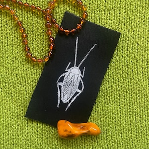 May include: A black fabric patch with a white embroidered image of a cockroach. The patch is on a green knitted surface with a brown amber necklace and a small orange stone.