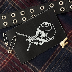 May include: A rectangular black fabric patch featuring a white embroidered graphic of a snail with a skull-shaped shell. The patch is accompanied by two silver-toned metal safety pins and a small, off-white tooth-like object.