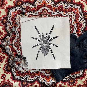 May include: A black and white printed image of a spider on a white canvas with a brown and white paisley patterned fabric background.