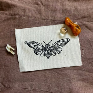 May include: A black and white printed design of a moth on a white canvas fabric. The fabric is on a brown surface.