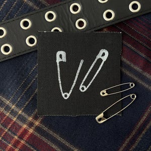 May include: A square black fabric patch featuring an embroidered white outline of two open safety pins. Two metallic silver safety pins are placed on the lower right corner of the patch, which is positioned near a black leather belt with metal eyelets.