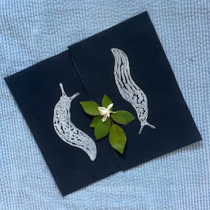May include: Two black cards with white ink illustrations of slugs and a white flower with green leaves.