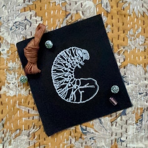 May include: A black fabric patch with a white embroidered image of a curled up grub or caterpillar.