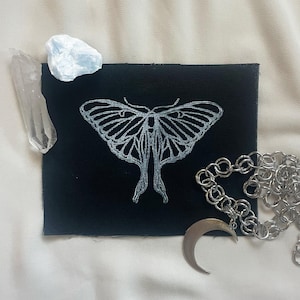 May include: A black fabric patch with a white outline of a moth with large eyes. The patch is surrounded by a silver chain and a crescent moon charm.