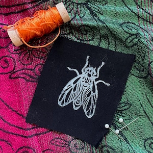 May include: A black fabric patch with a silver fly design, orange thread spool, and two pearl-headed pins. The patch is on a patterned fabric with pink and green hues. The fly design is detailed and stylized.