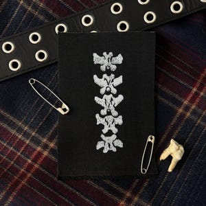 May include: A rectangular black fabric patch featuring a vertical white embroidered design of five stacked anatomical vertebrae. The patch is accompanied by two silver metal safety pins, a black leather belt with metal grommets, and a single tooth.