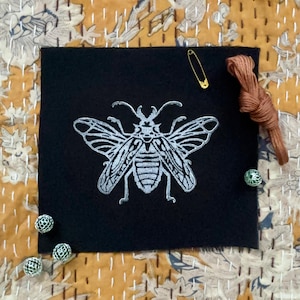 May include: A black fabric patch with a silver embroidered insect design. The insect has wings and a segmented body.