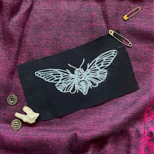 May include: A black fabric patch with a white outline of a moth with a wingspan of approximately 6 inches. The moth is centered on the patch and has a detailed design.